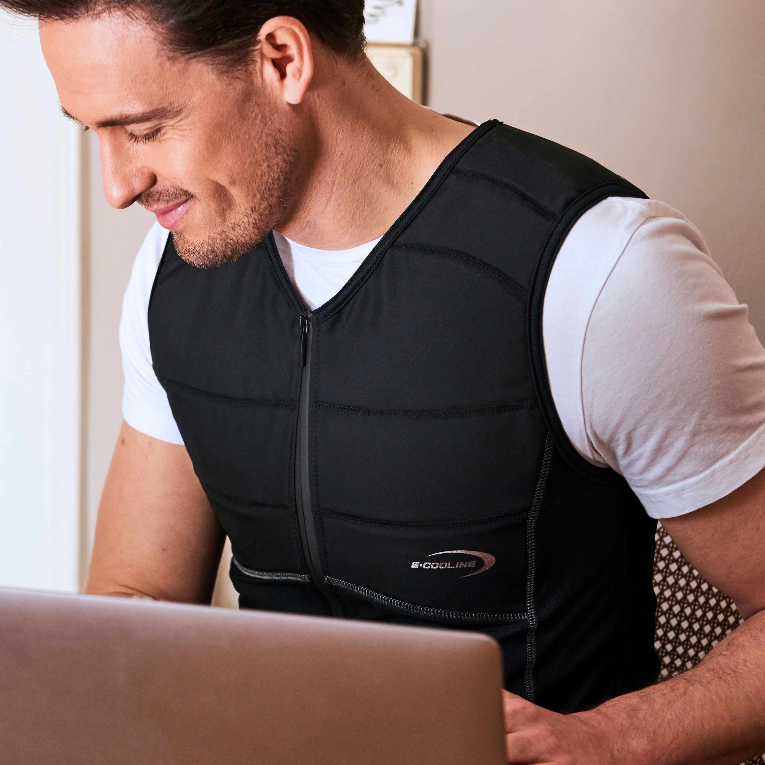 E.COOLINE Powercool SX3 - maglia smanicata refrigerante - il condizionatore da indossare Uomo indossa una maglia smanicata refrigerante E.COOLINE di colore nero e lavora al suo computer portatile.
